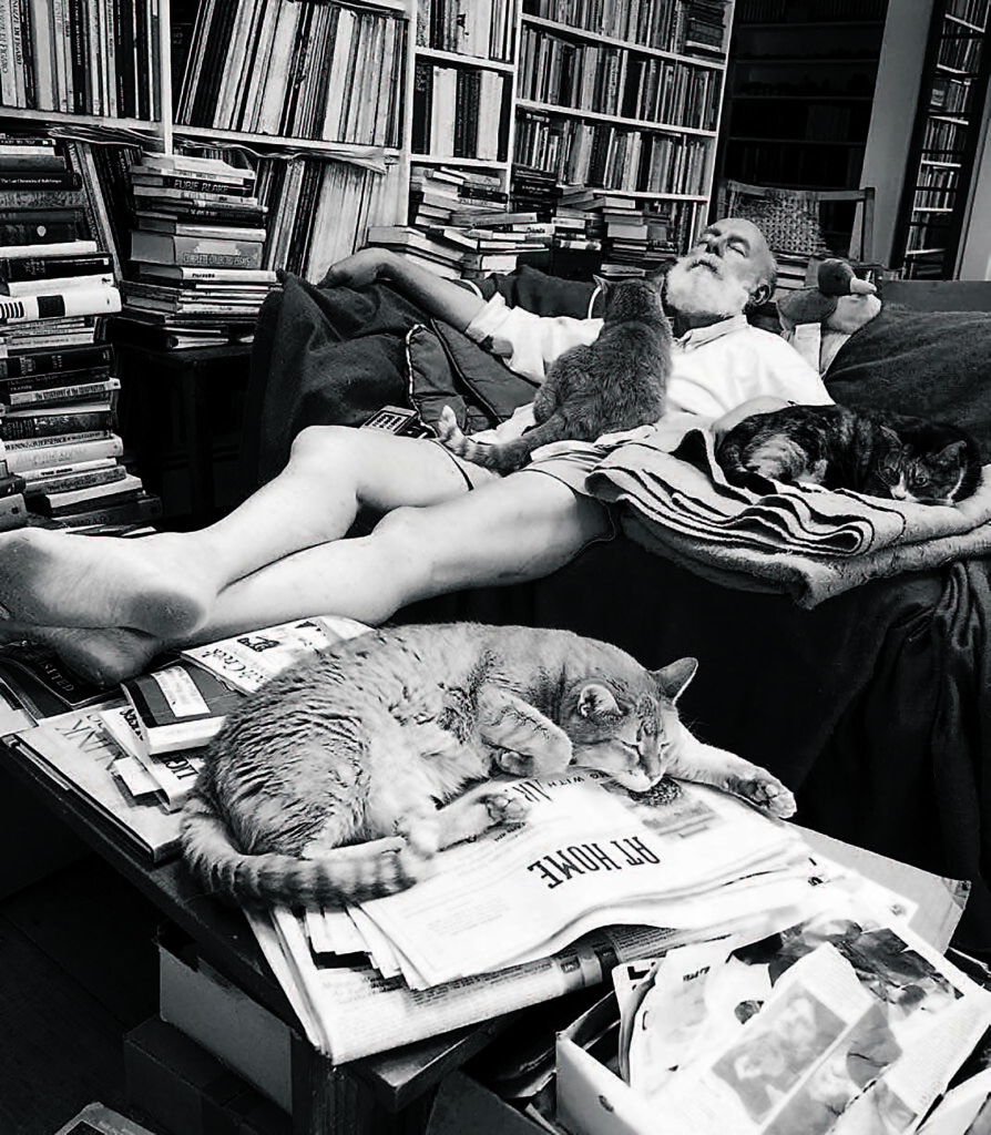 a man lounging in a recliner sleeps while his three cats join him by napping around him