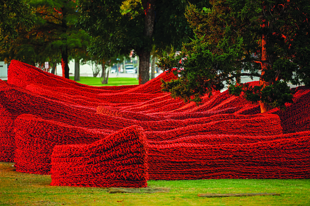 Red woven textile installation creates undulating, landscape-like shapes across a lawn, with mature trees rising through the artwork