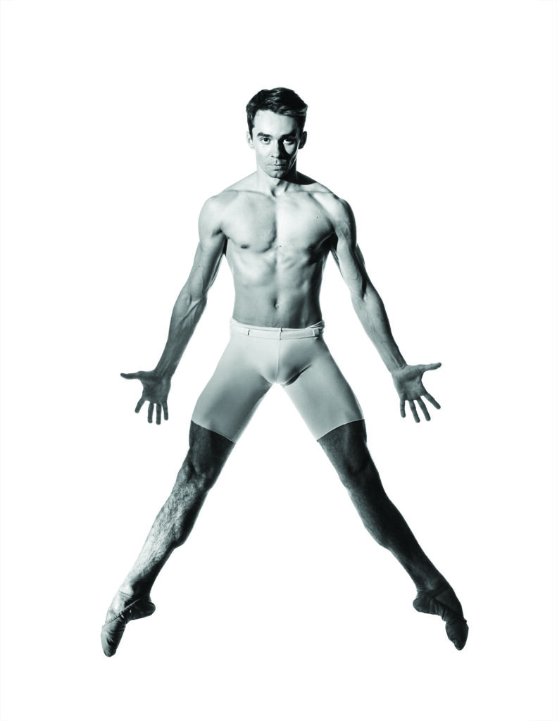 A man in tight white shorts jumps up against a bright white background while dancing