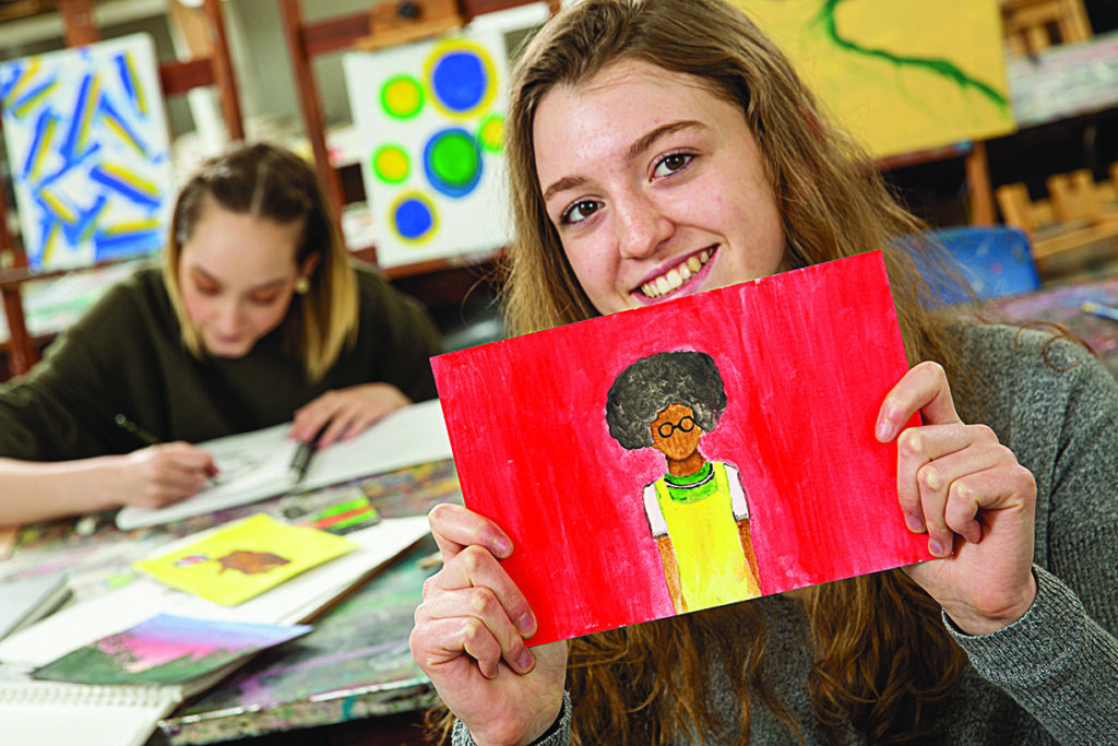 An student artist holds up a brightly colored portrait while seated in a classroom at a table while another student continues to create art at the table across from them