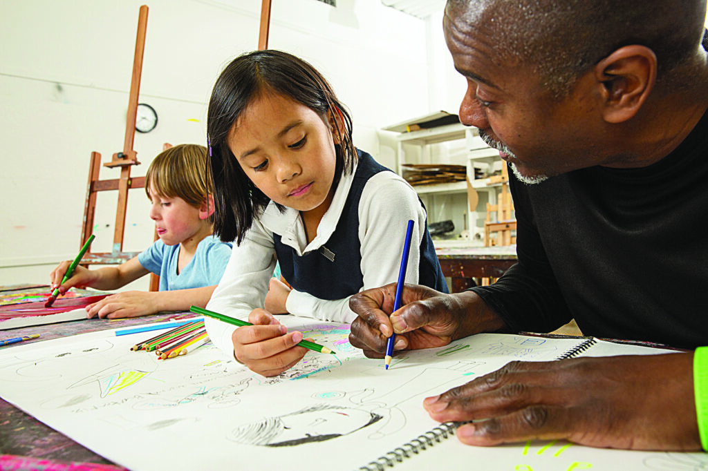 An adult instructor guides two children as they draw at a table covered with sketchbooks and colored pencils