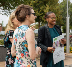 three women stand facing right, smiling and in conversation in an outdoor setting