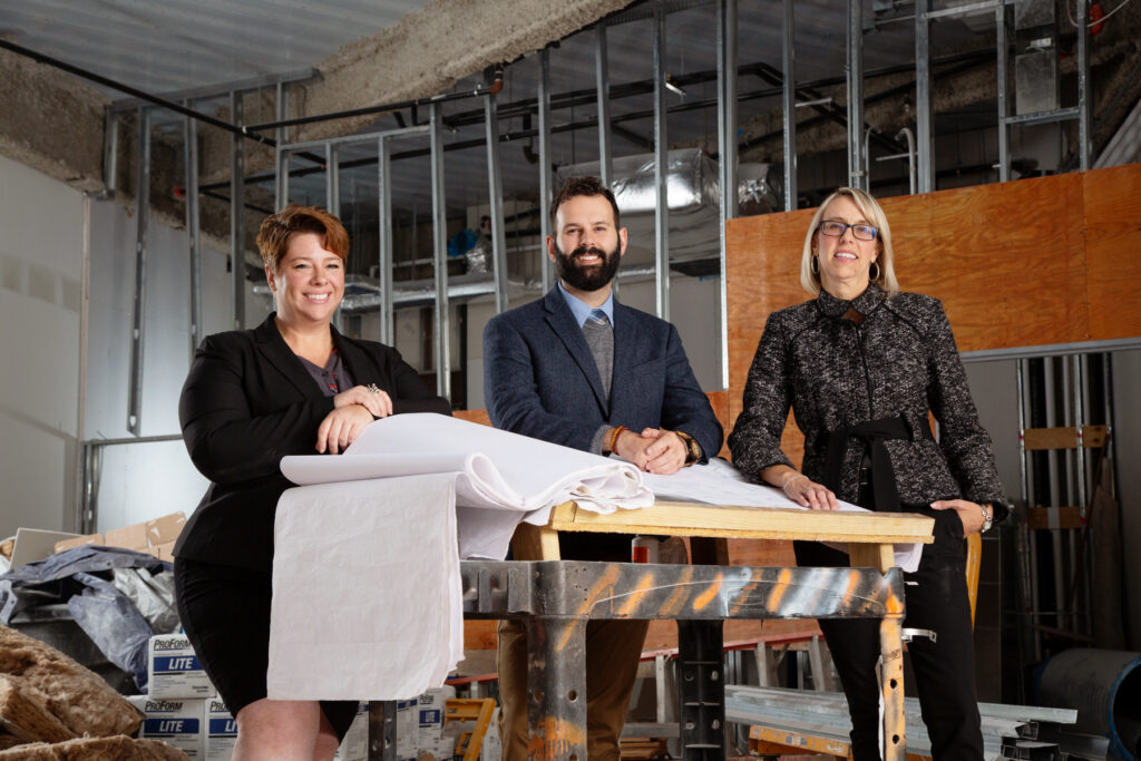 Three adults in professional dress stand behind an architect's desk in an industrial setting