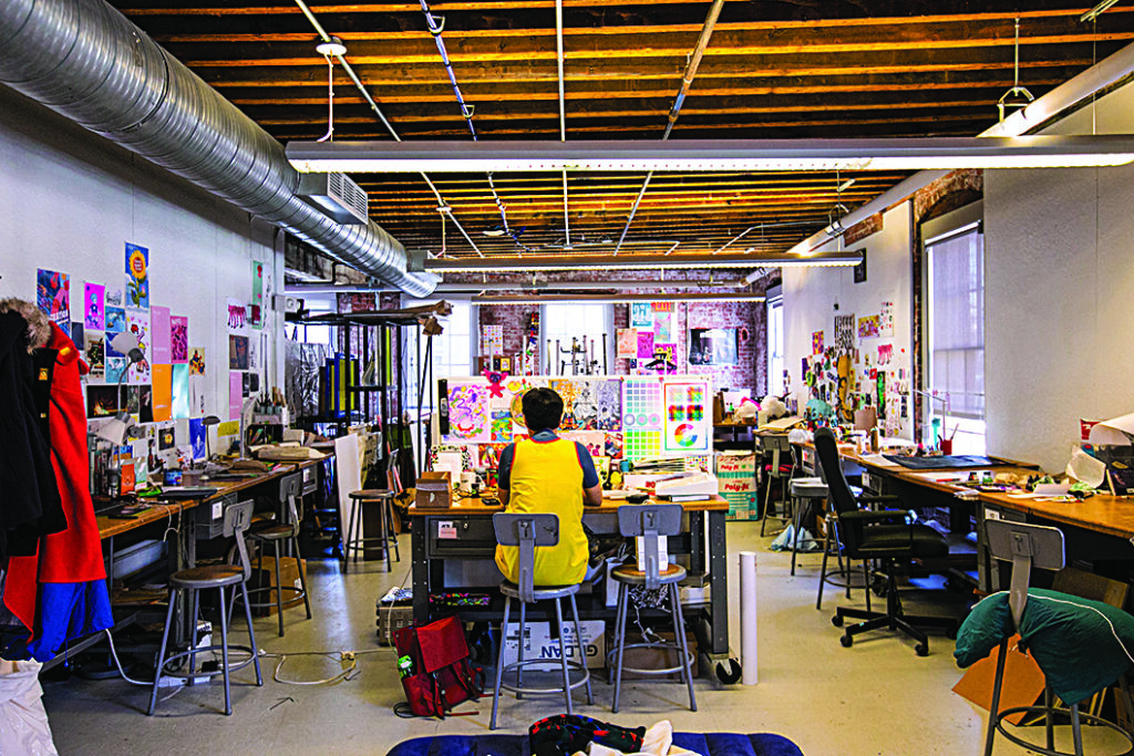 Person seated at a worktable in a large open art studio, surrounded by desks, tools, colorful artwork, and posters displayed on the walls.