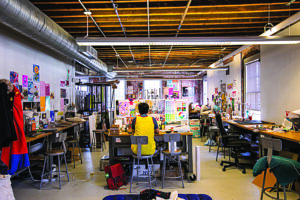 Person seated at a worktable in a large open art studio, surrounded by desks, tools, colorful artwork, and posters displayed on the walls.