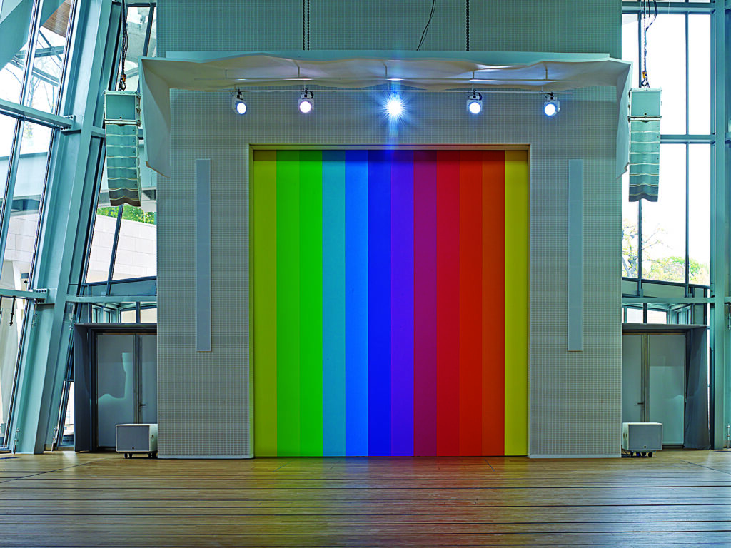 Contemporary art installation consisting of tall vertical panels in a spectrum of colors, from green and blue to purple, red, and yellow, set within a modern glass-and-steel interior. Overhead spotlights illuminate the center of the artwork, with speakers and architectural elements framing the installation.