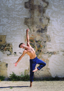 a man dances barefoot in the sand in front of a rustic wall while wearing blue pants in an outdoor setting