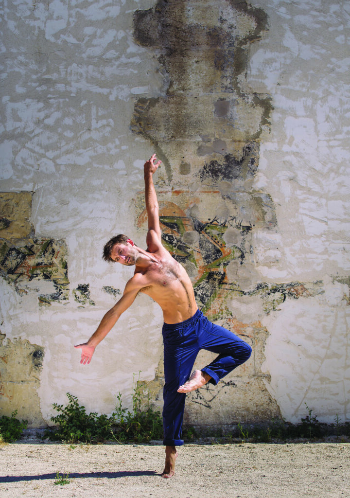 a man dances barefoot in the sand in front of a rustic wall while wearing blue pants in an outdoor setting