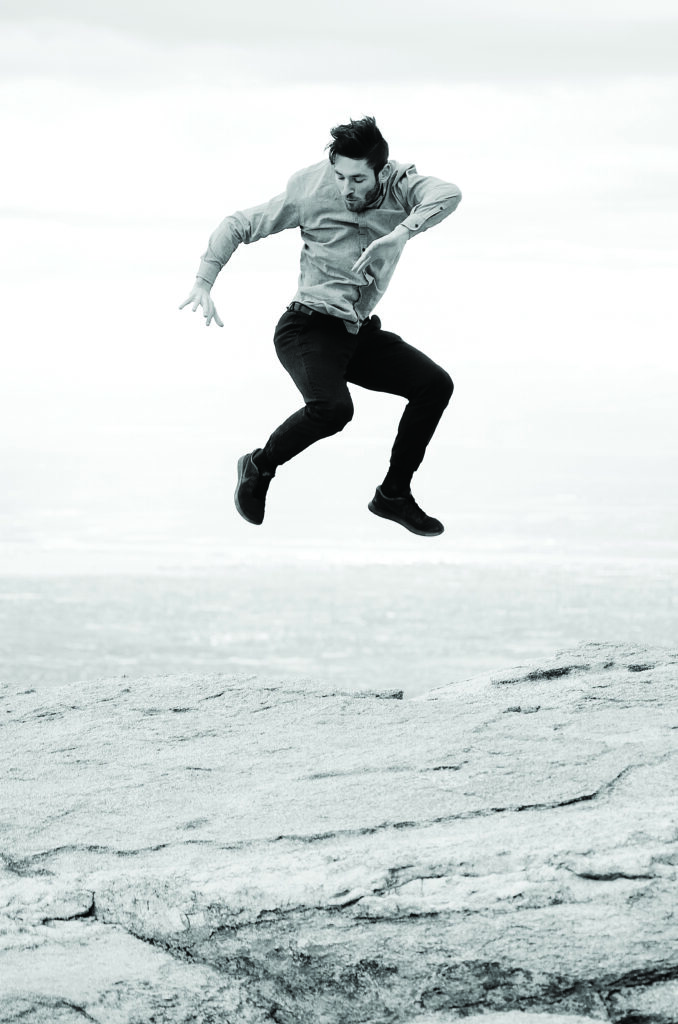 A man jumps in a dress shirt and pants on a rock above an ocean in a dramatic dance performance