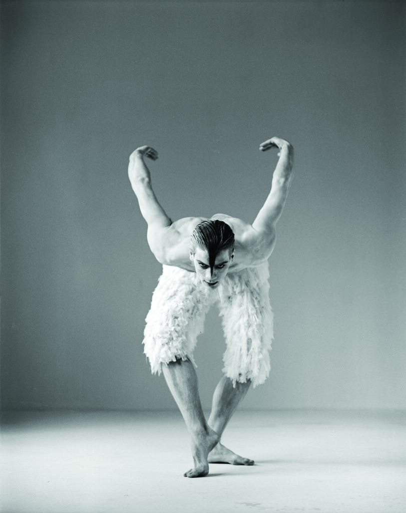 Black-and-white studio photograph showing a ballet dancer balancing with crossed legs and a forward-leaning torso. The dancer's arms curve upward on either side of the body, and a textured, feather-like costume covers the lower half. The background is plain, drawing attention to the dramatic pose and body lines.