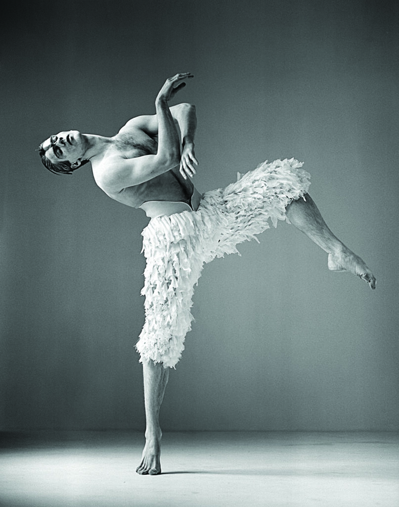 Black-and-white studio photograph showing a ballet dancer in a textured, feather-like costume. The background is plain, drawing attention to the dramatic pose and body lines.