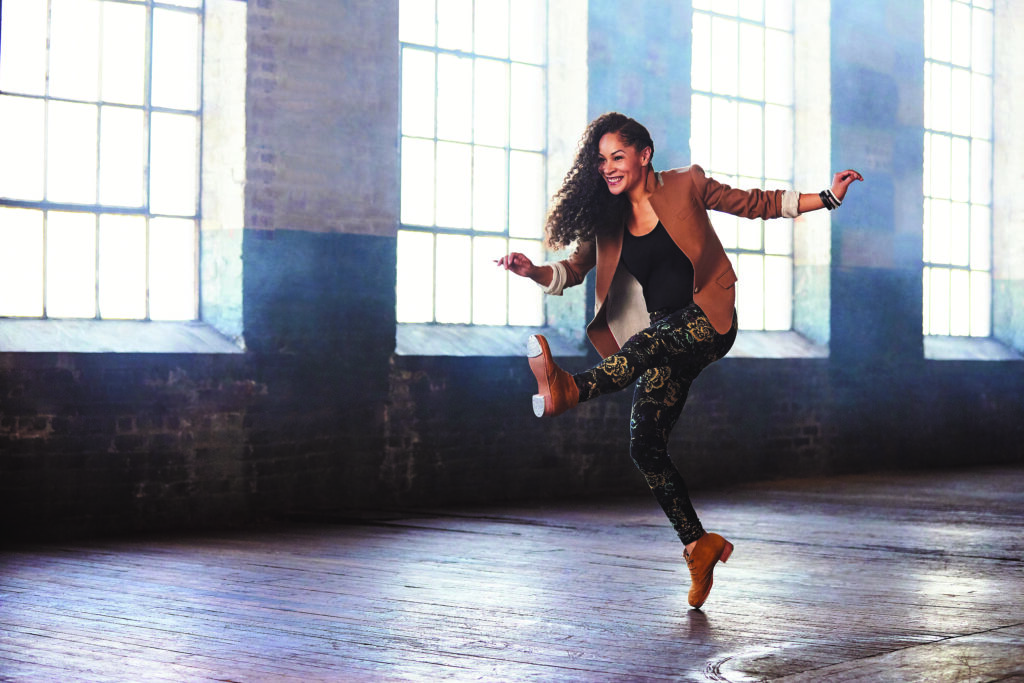 Acclaimed tap dancer Ayodele Casel lifts one leg high while dancing in a studio, exposing her tap shoe plates and smiling during her performance