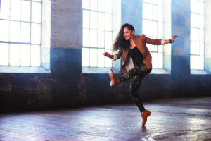 Acclaimed tap dancer Ayodele Casel lifts one leg high while dancing in a studio, exposing her tap shoe plates and smiling during her performance