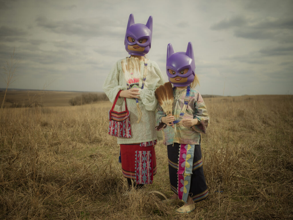 Two women in traditional dress stand in a field while wearing Lego heads with purple Batman masks