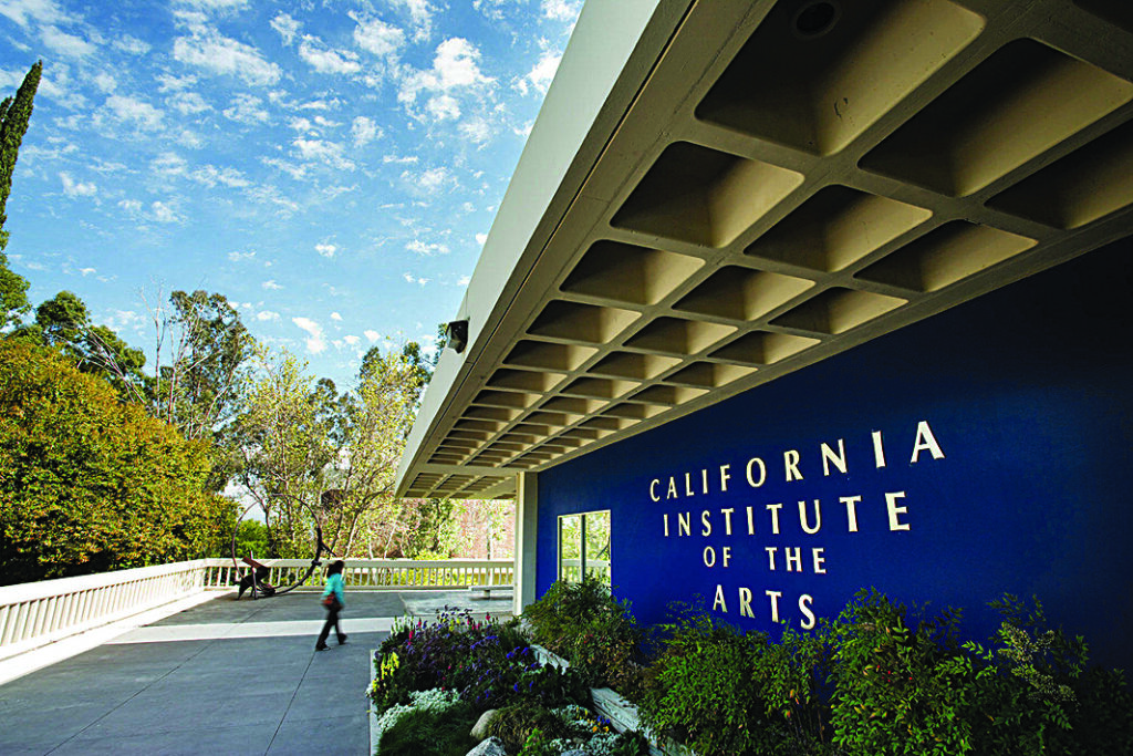 Exterior of the California Institute of the Arts building, with the school's name displayed on a blue wall beside a landscaped walkway bordered by trees and shrubs.