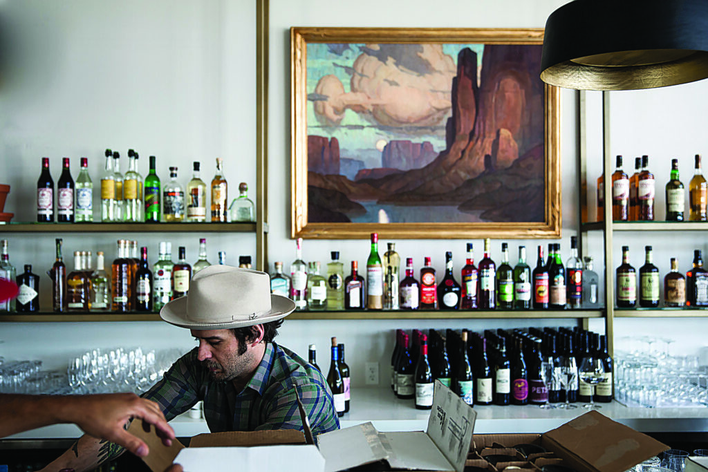 Person wearing a light-colored hat working behind a bar counter, with shelves of liquor bottles and a large, framed landscape painting on the wall behind.