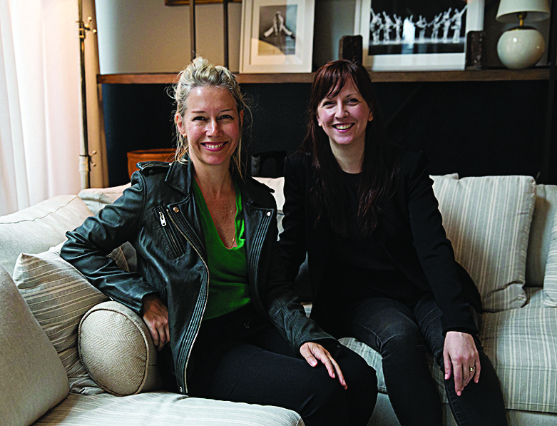 Two women in modern fashionable clothes, Sarah Crowner and Jessica Lang, sit together on a cream-colored couch and pose for a photo