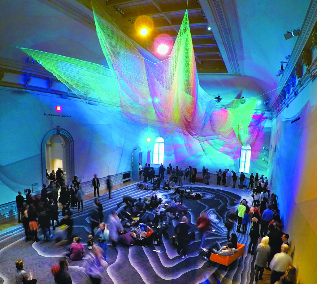 Large multicolored net installation suspended from the ceiling of a gallery, above a floor patterned with flowing contours and a crowd of visitors gathered throughout the space.