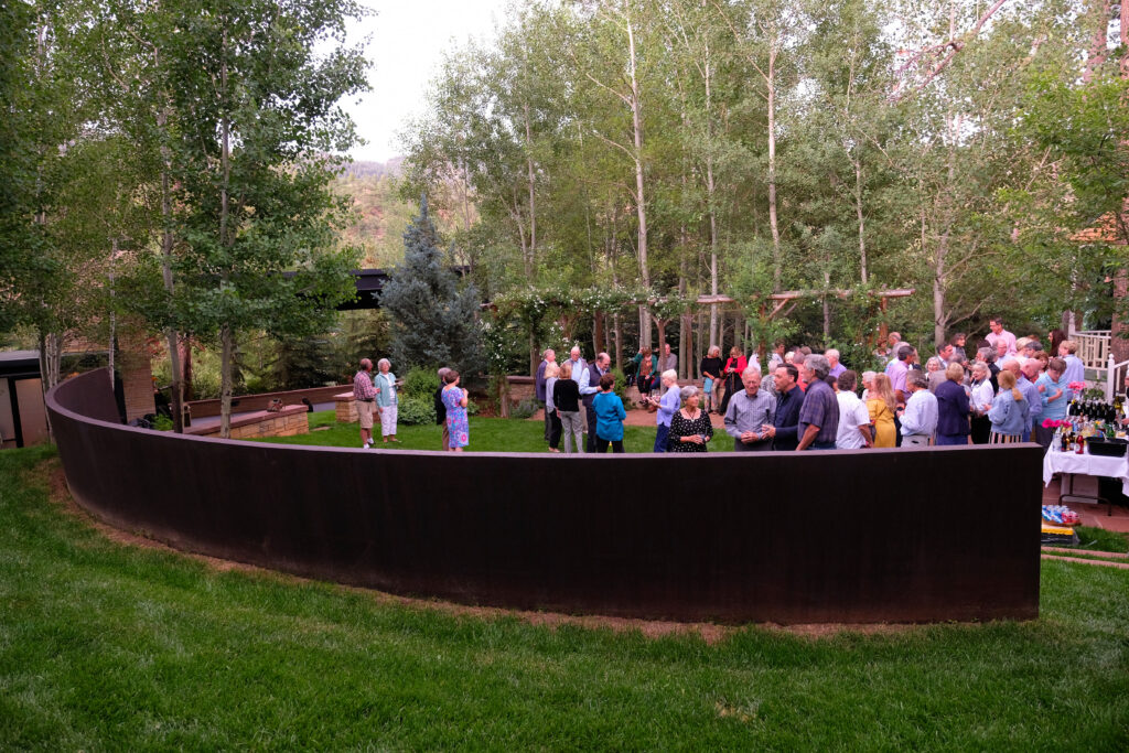 A large outdoor gathering in a landscaped garden where people stand and talk near a curved metal wall