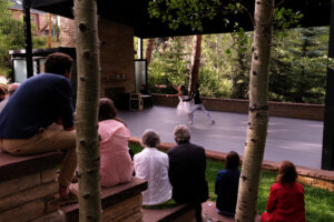An outdoor ballet performance on a forest‑side stage, with dancers performing before a seated audience