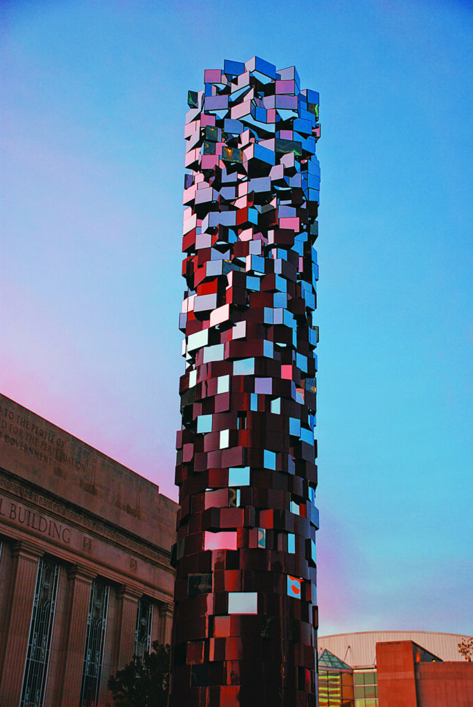 A tall vertical sculpture made of hundreds of reflective rectangular blocks in varying sizes and shades of red, white, blue, and dark gray. The blocks create a fragmented, geometric appearance as the structure extends high into a clear blue sky. A stone building is visible on the left, with additional modern buildings in the background.