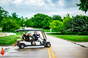 a golf cart full of people crosses a lush green tree-lined street