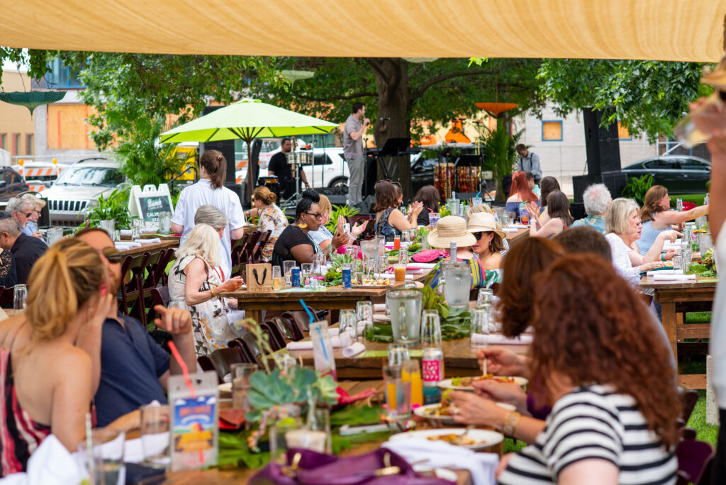 A lively outdoor brunch event where people sit at long tables under a canopy, sharing food and conversation