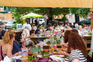 A lively outdoor brunch event where people sit at long tables under a canopy, sharing food and conversation