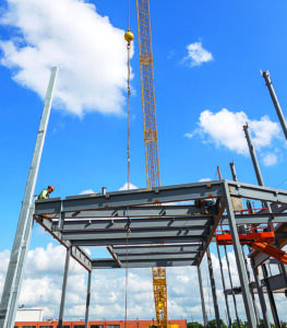 A steel building frame under construction with workers securing beams beside a large crane