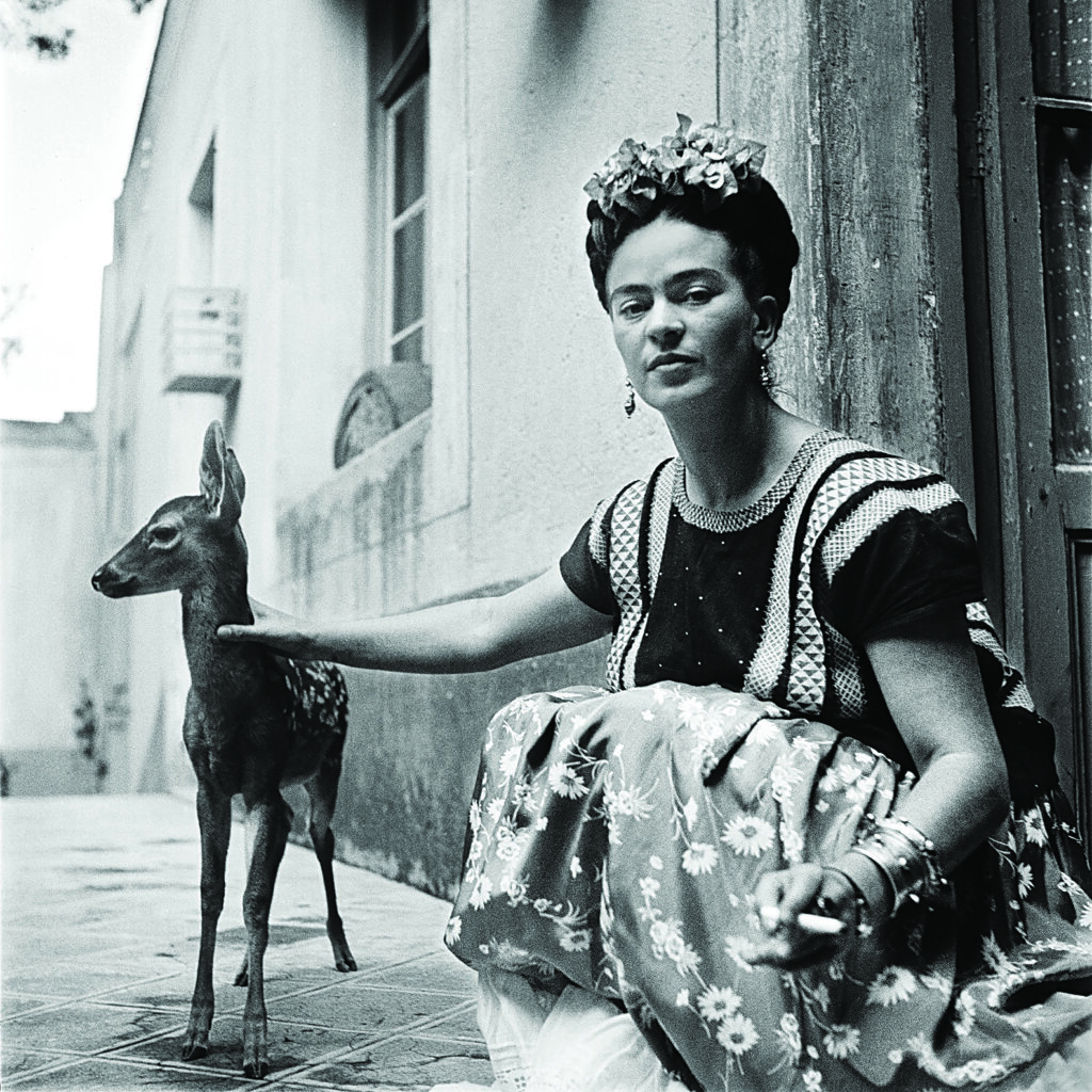 a woman in traditional Mexican dress kneels outside a building and pets her fawn