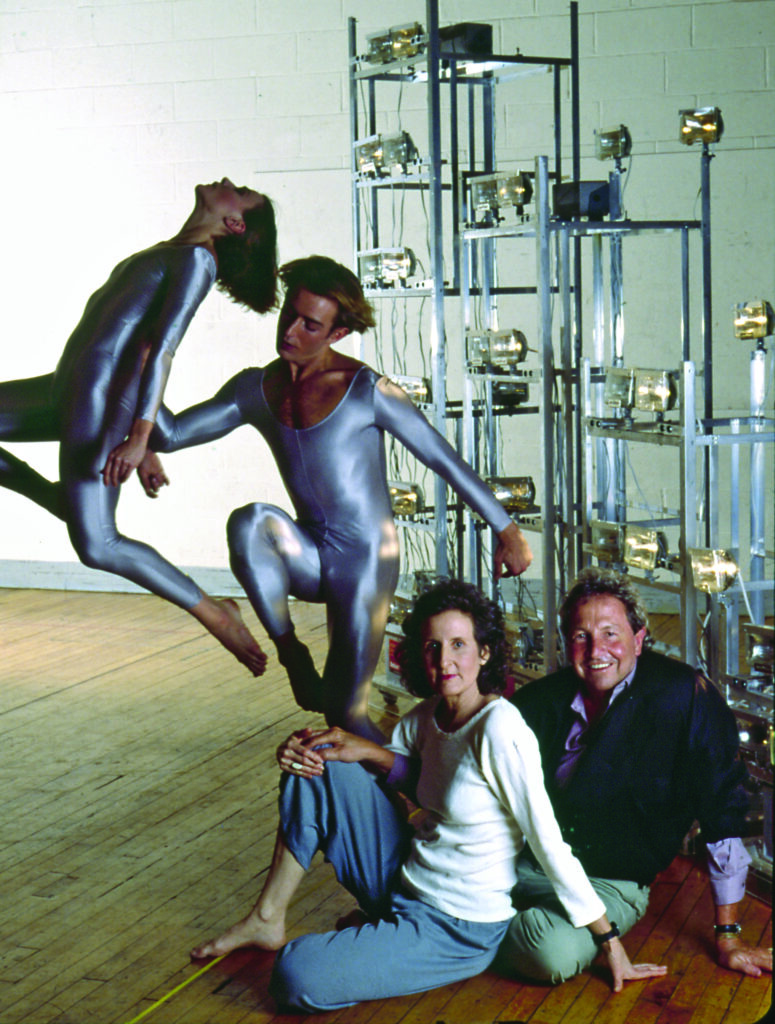an artist and choreographer sit on the hardwood floor of a dance studio while performers in silver leotards dance behind them next to an industrial lighted shelfing wall