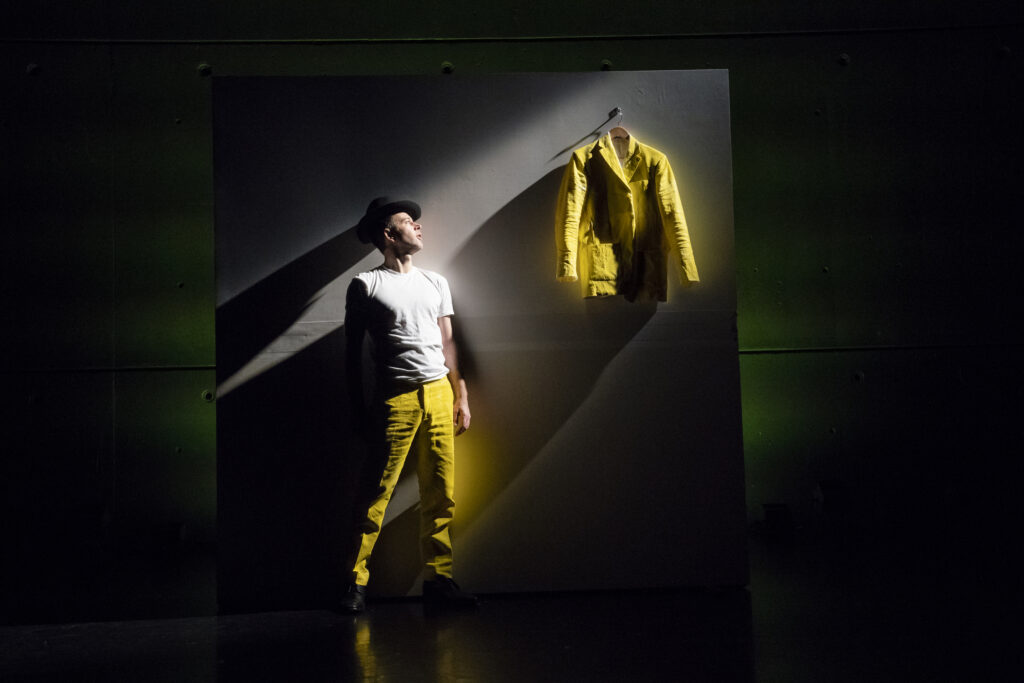 A dancer in a yellow suit stands before a dramatically lit wall with their removed yellow jacket hanging in a spotlight