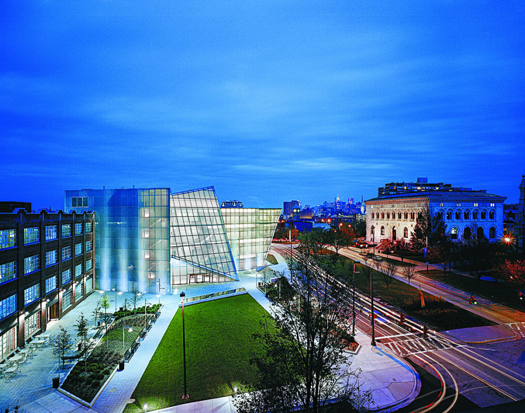 Contemporary glass academic building illuminated at dusk beside a landscaped lawn and city street, with light trails from passing vehicles and additional campus buildings in the background.