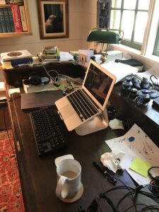 A cluttered wooden desk with an open laptop, scattered papers, books, and office supplies showing a lived-in and creative work space.