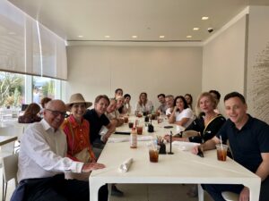 a group of people gathered together, seated around a table for a lunch look at a camera at one end of the table and smile