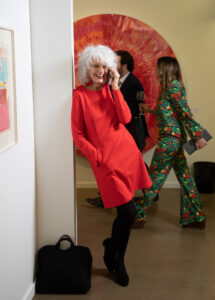 a woman in a vibrant red coat leans against a wall in an art gallery