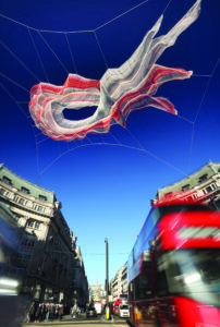 Red and white net sculpture suspended above a city street between historic buildings, with red double-decker buses and traffic passing below under a clear blue sky.