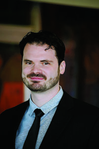 Portrait of Jeremiah Davis, who stands in an art gallery in a suit and tie as he smiles