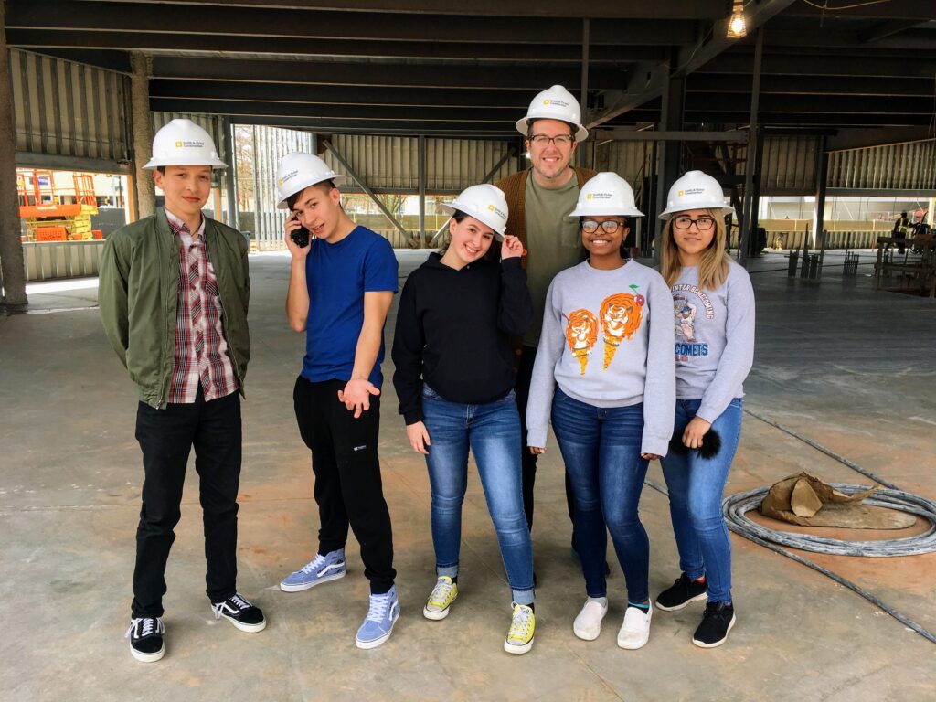 a group of teenagers and their adult advisor wear hardhats to visit a construction site