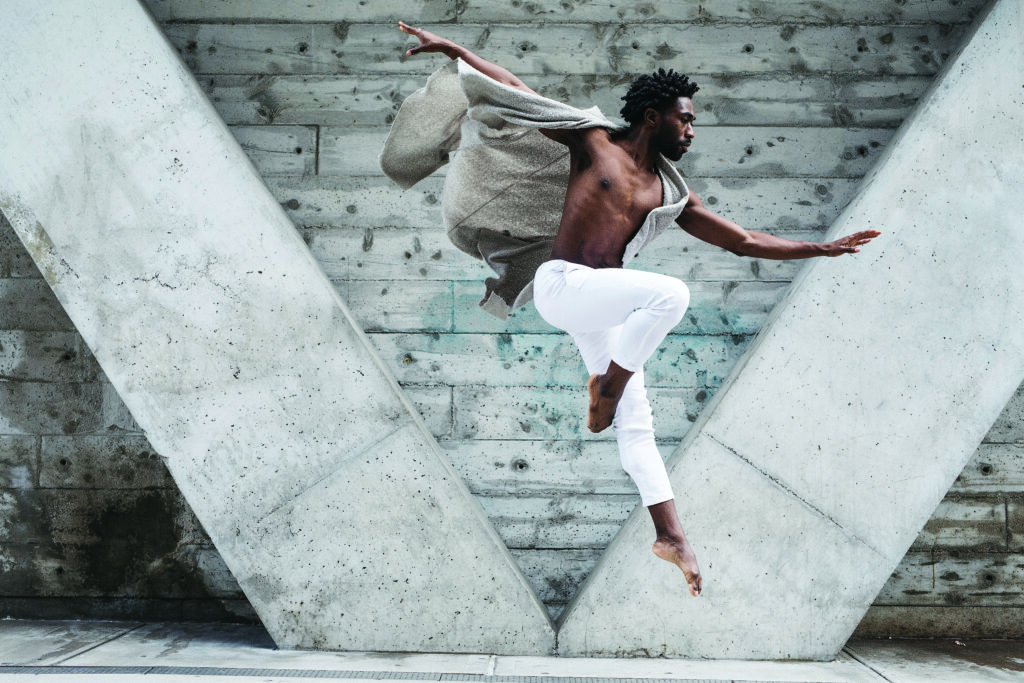 a man in a loose grey vest and tight white pants leaps to the right while dancing in an industrial setting