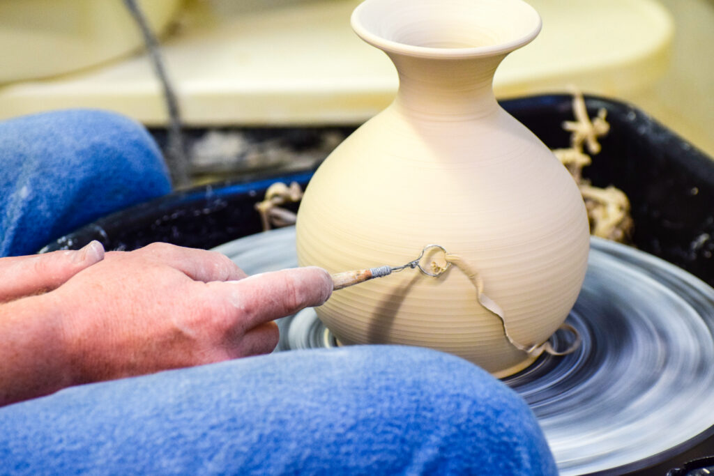 A hand uses a tool to create decorative texture on a clay pot on a pottery wheel