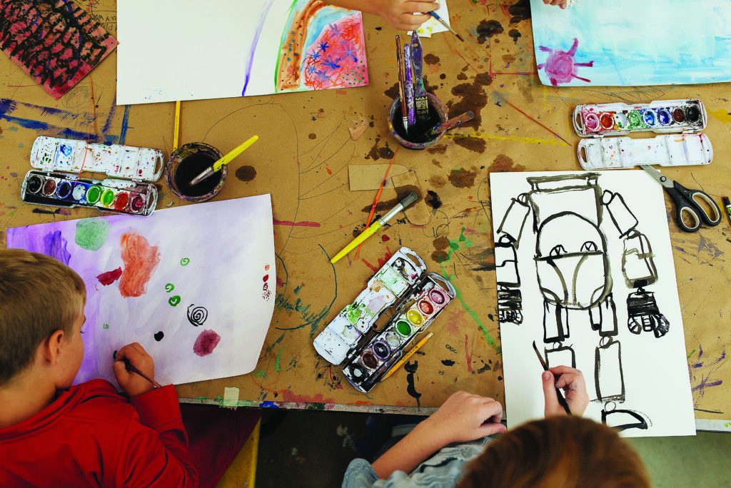 A brown-paper wrapped tabletop is covered with pencils, paint palettes, brushes, and colorful works‑in‑progress as two children explore watercolor