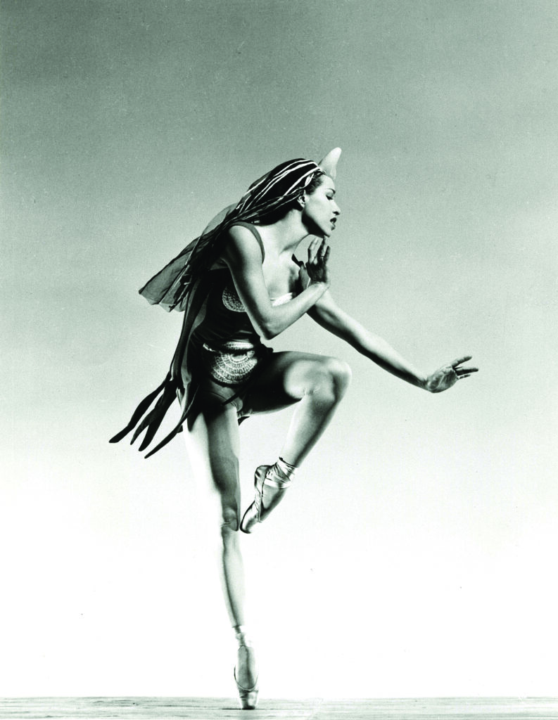 A prima ballerina on point leans to the right while pulling one up so her toe touches her thigh in an artistic costume designed by Isamu Noguchi
