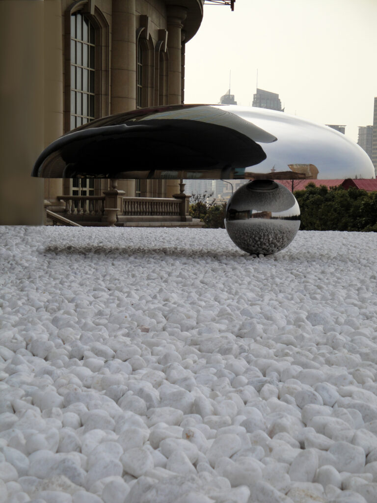 A reflective metal dome balanced on a reflective metal sphere in on a bed of white gravel in an outdoor setting in front of a building facade