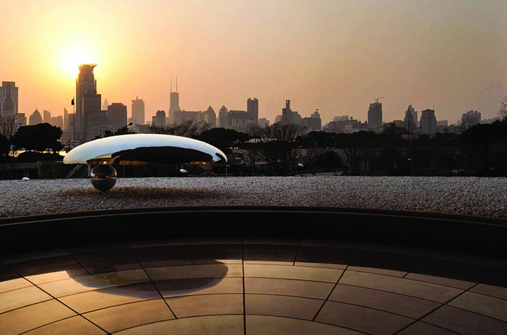 A reflective metal d sculpture set beneath a shaded structure with a sunset skyline in the background