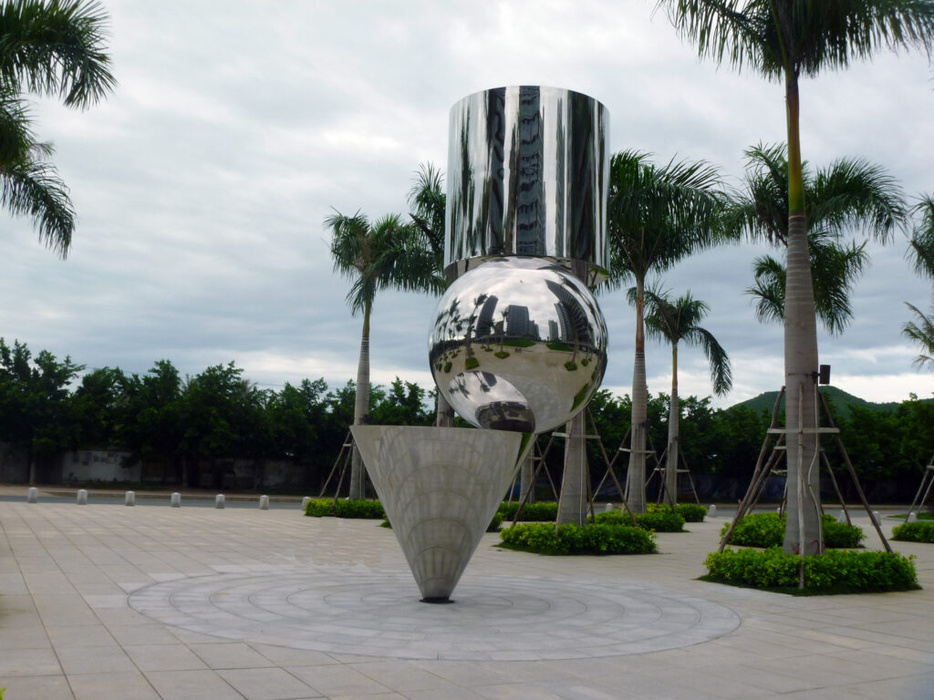 A tall outdoor sculpture made of stacked reflective geometric shapes—cone, sphere, and cylinder— in a plaza flanked by palm trees