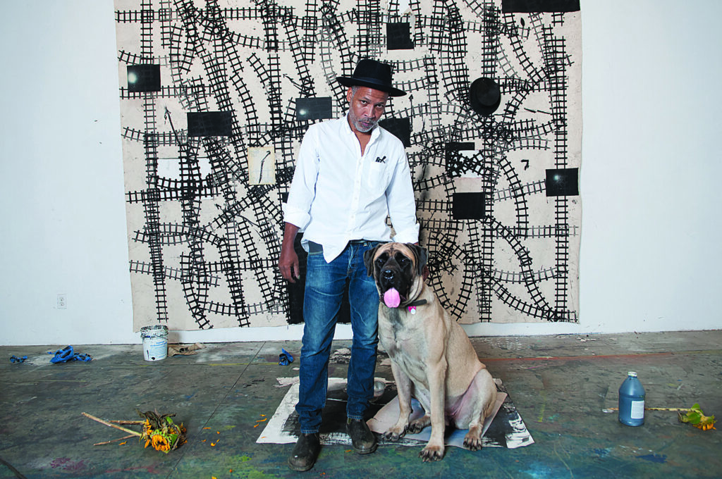 A man in a casual button down and jeans wears a hat while his dog sit beside him in front of an artwork in a studio