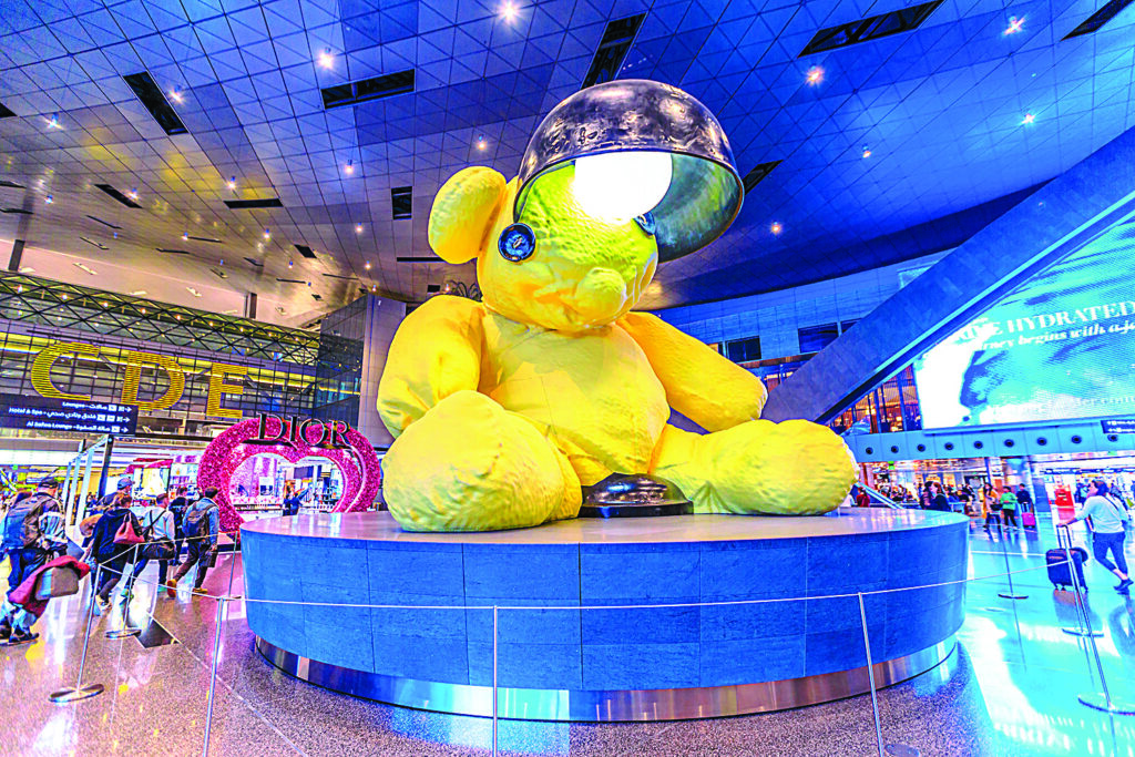 a stuffed yellow bear sculpture with button eyes sits on display at an airport