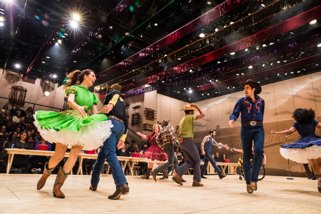 The cast of "Oklahoma!" on Broadway give a lively stage performance in Western‑style costumes moving energetically on a decorated wooden stage