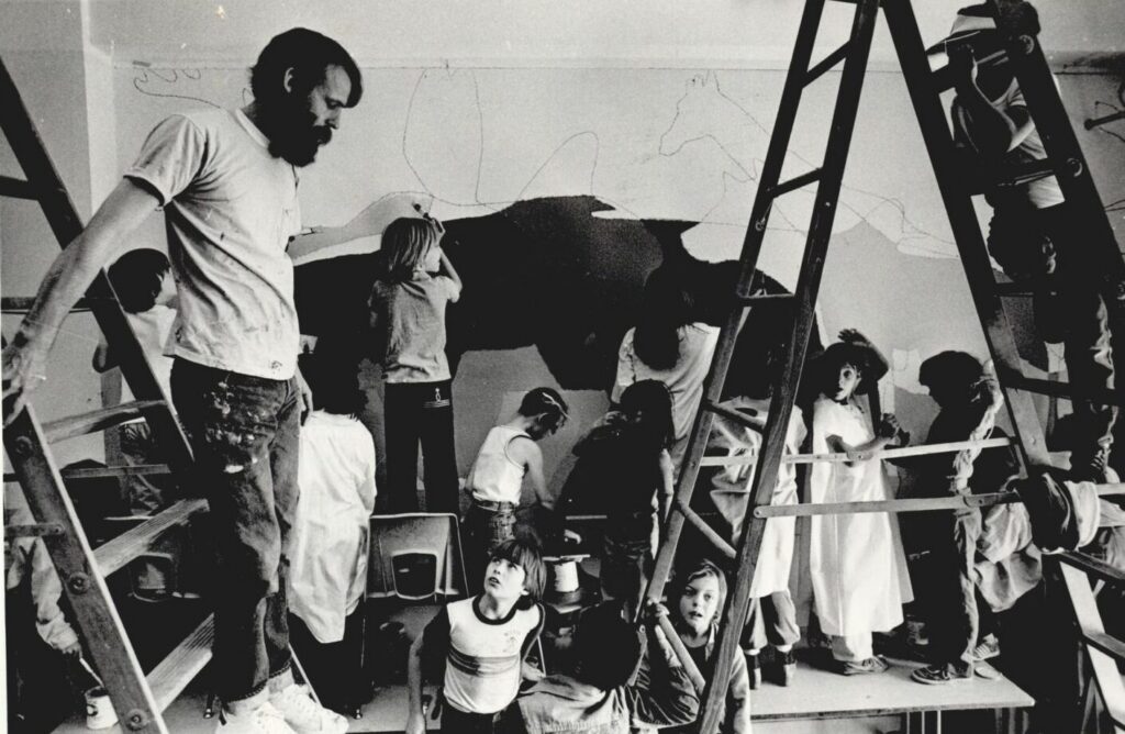 A artist, surrounded by elementary school students, oversees the painting of a mural at an elementary school in 1980